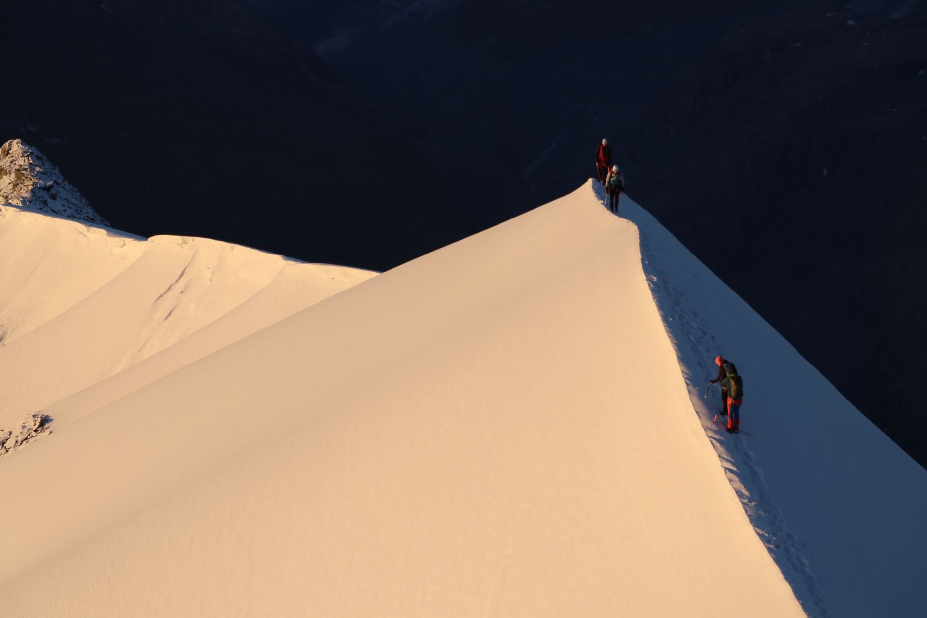 Alpinistes progressant sur une arête enneigée éclairée par une lumière dorée, au-dessus d’une vallée sombre.