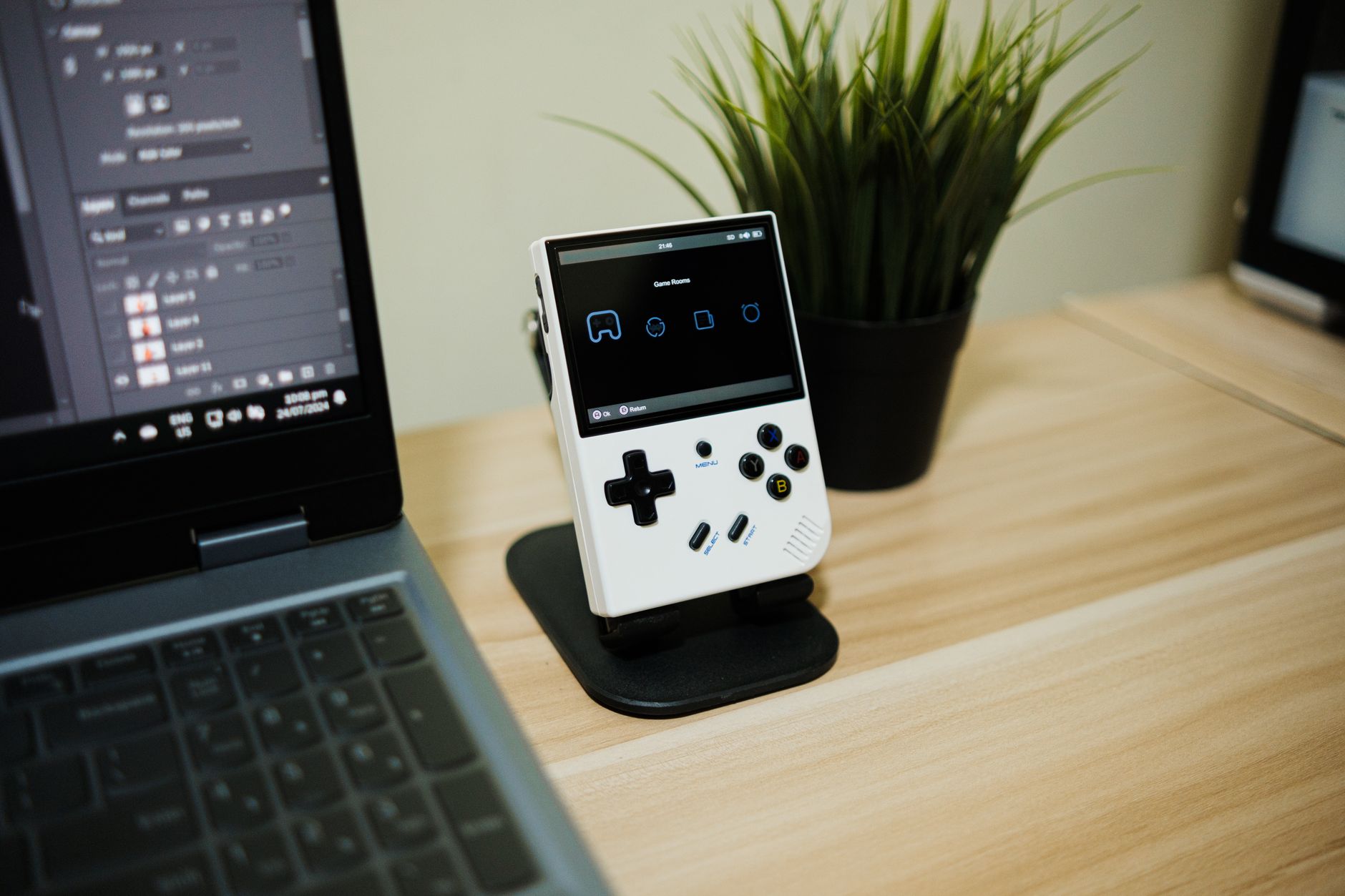 Console portable rétro blanche sur un support noir, posée sur un bureau en bois près d’un ordinateur portable et d’une plante.