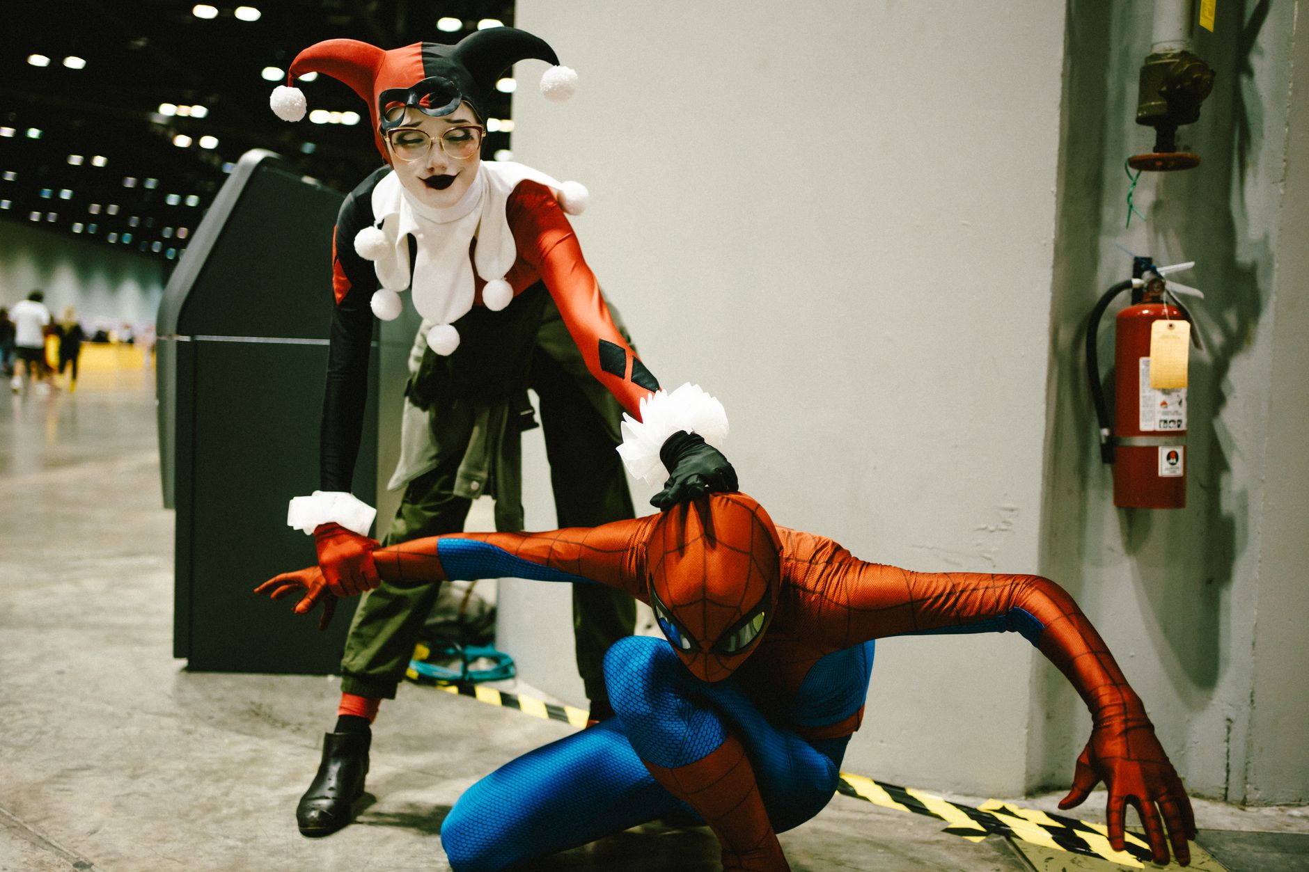 Deux cosplayeurs posent dans un hall : l’un en costume d’arlequin rouge et noir, l’autre accroupi en costume de héros rouge et bleu.