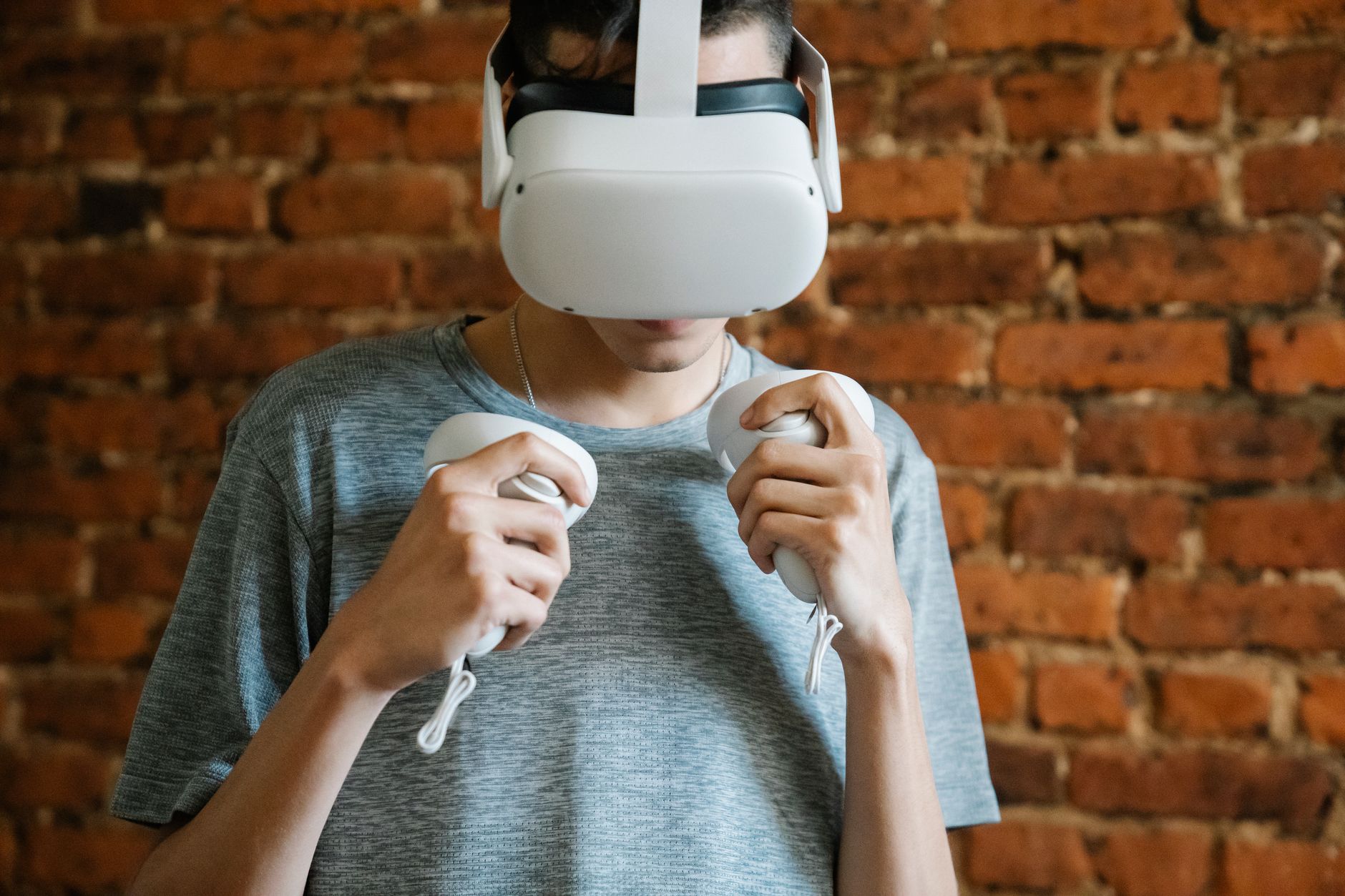 Personne en t-shirt gris portant un casque de réalité virtuelle blanc et tenant deux manettes devant un mur de briques rouges.