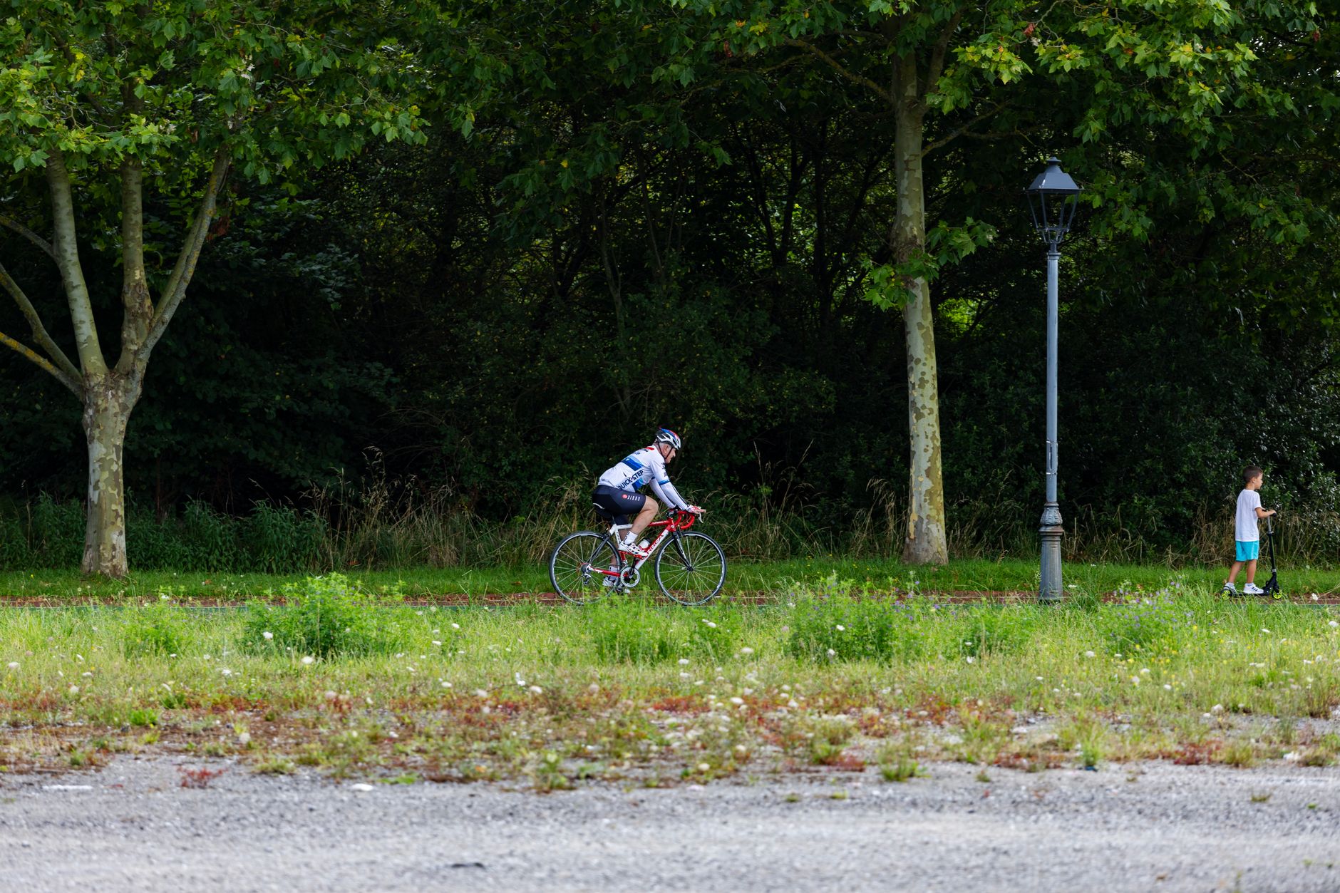 Un homme équipé d’un casque roule à vélo près d’arbres, tandis qu’un enfant se déplace en trottinette à côté.