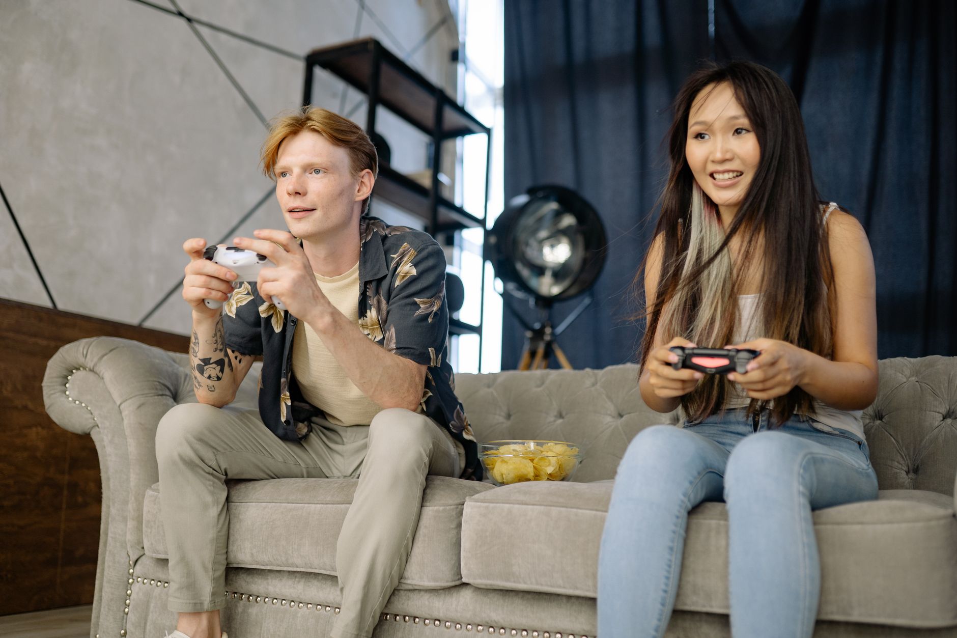 Un homme et une femme assis sur un canapé gris tiennent chacun une manette de jeu vidéo, avec un bol de chips posé entre eux.